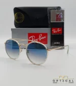 Lunettes de soleil Ray-Ban Round Double Bridge RB3647N dorées avec cerclages en acétate transparent et verres dégradés bleus, étui noir, boîte grise et livret de garantie chez Optical Gallery.
